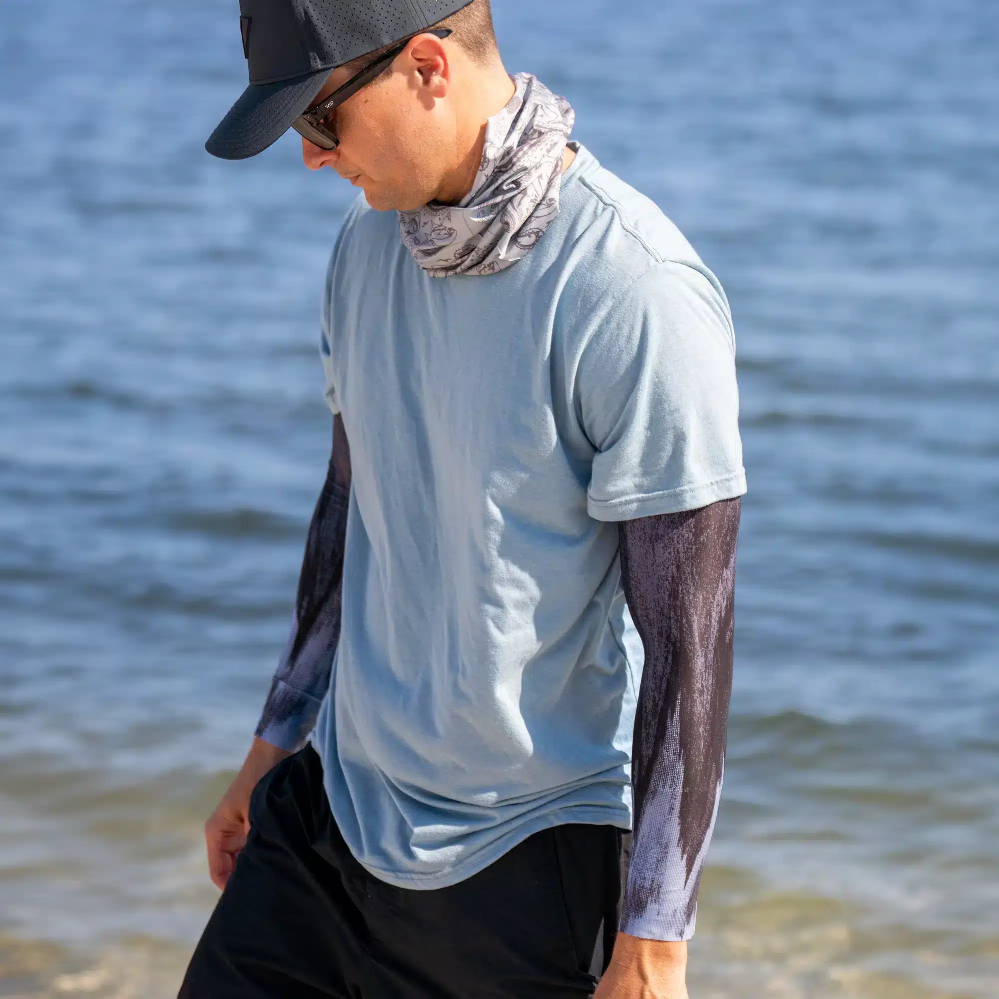 An image of a person standing near the ocean. They are in a navy blue hat, sunglasses, a paisley neck gaiter, a light blue shirt, and a matching pair of arm sleeves.