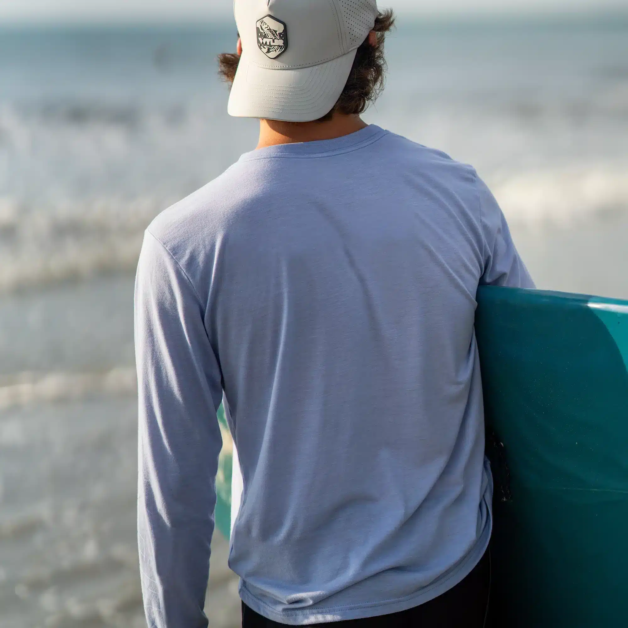 An image of a person holding a surfboard and walking toward the ocean. They are wearing an off-white hat and a light blue shirt. The hat features a tarpon breaching water.