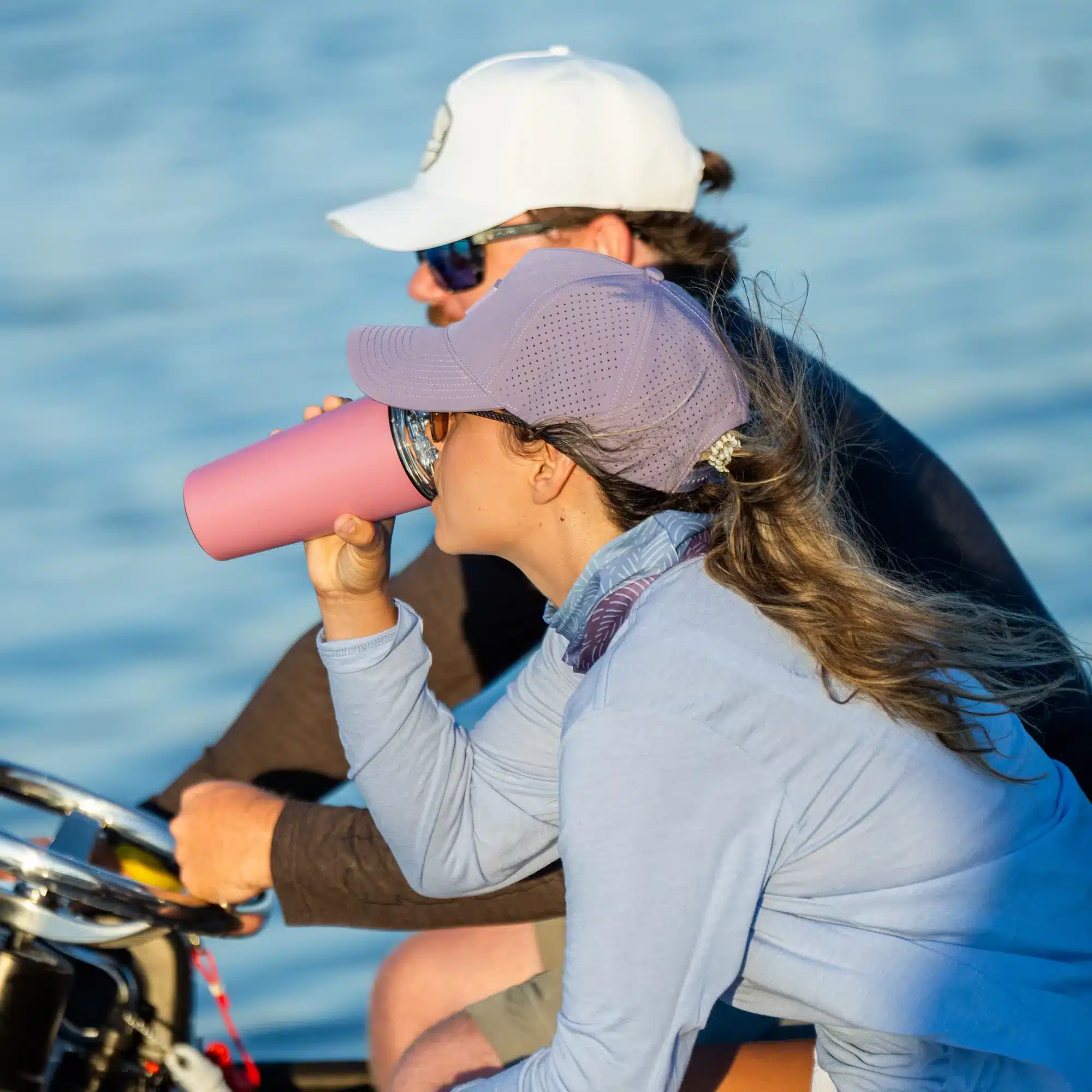 an image of two people in a boat. The one driving it is in a white hat. The passenger is in a purple hat and drinking from a pink tumbler.