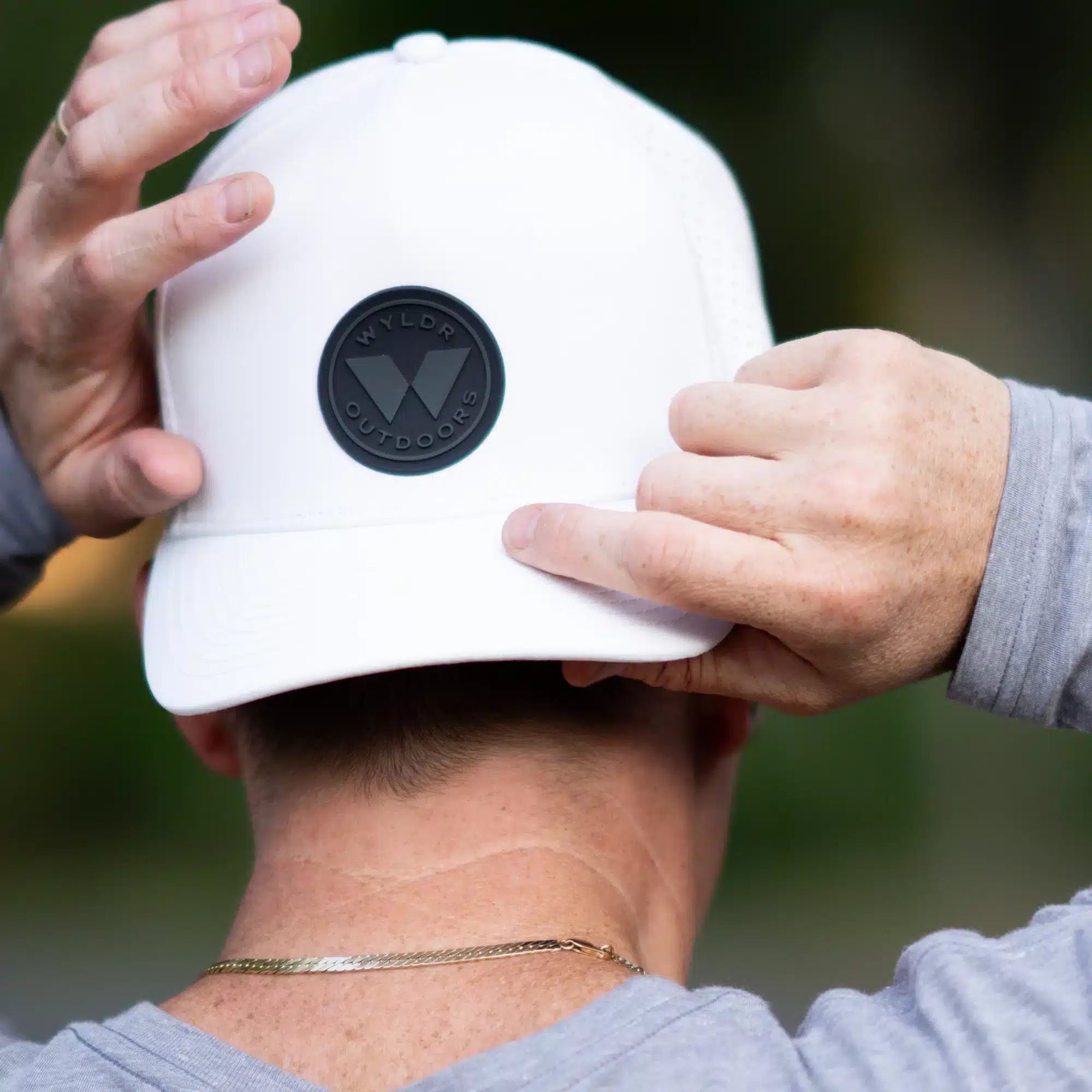 An image of a person putting on an off-white hat. It has a round silicone patch. The text reads Wyldr Outdoors.
