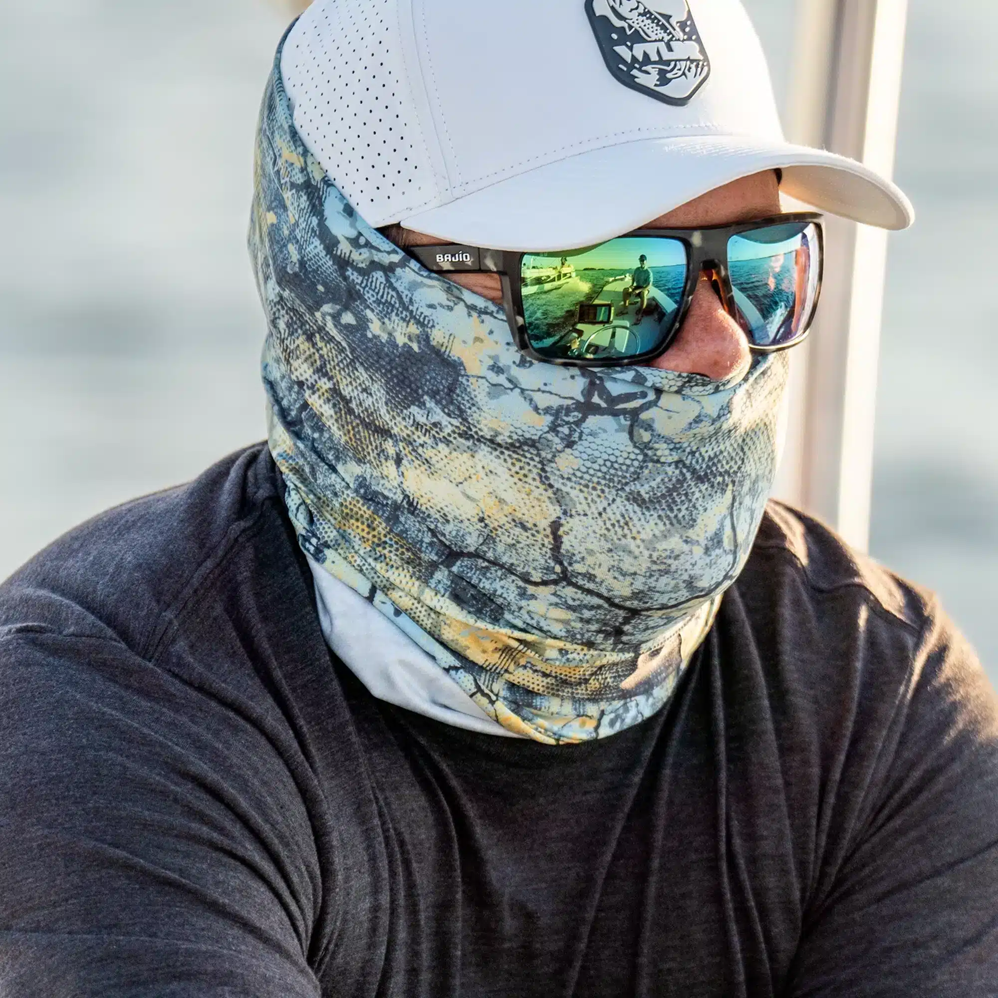 An image of a person driving a boat. They are in an off-white colored hat, a neck gaiter, sunglasses, and a charcoal black shirt. The hat features a tarpon fish breaching water. You can see the passengers in the reflection of the sunglasses.