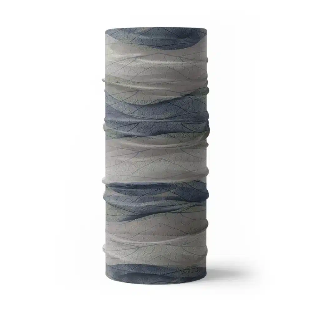 an image of a full neck gaiter. The design features lines and breaks to recreate the look of a leaf print. The colors behind the print mimic waves. This image showcases colors in blues and grays.