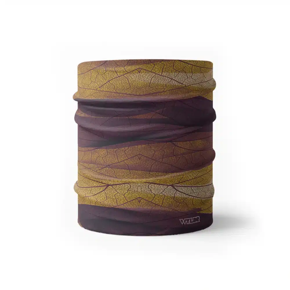 an image of a half neck gaiter. The design features lines and breaks to recreate the look of a leaf print. The colors behind the print mimic waves. This image showcases colors in warm purples, oranges and yellows.