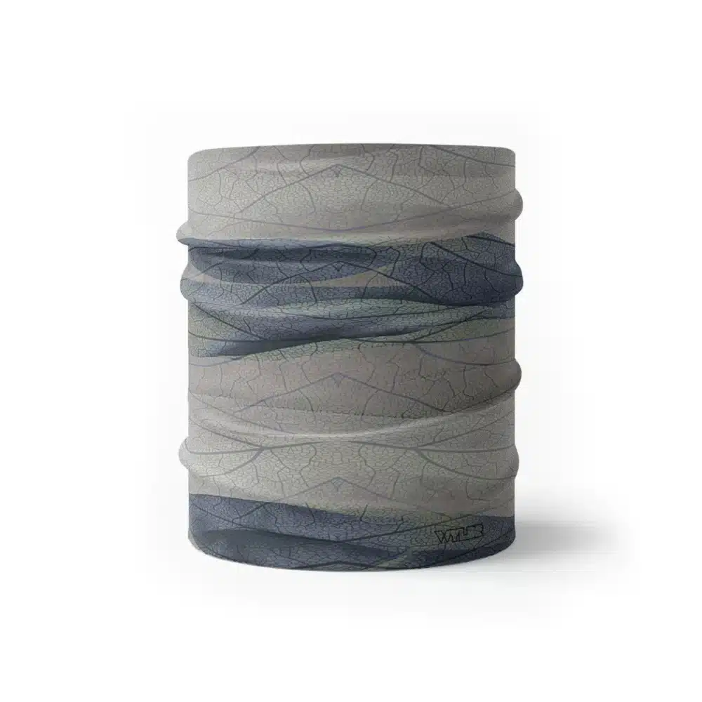 an image of a half neck gaiter. The design features lines and breaks to recreate the look of a leaf print. The colors behind the print mimic waves. This image showcases colors in cool blues and grays.
