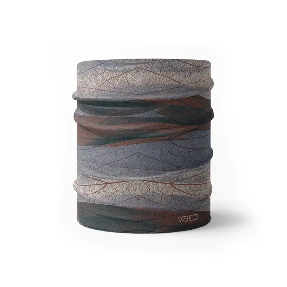 an image of a half neck gaiter. The design features lines and breaks to recreate the look of a leaf print. The colors behind the print mimic waves. This image showcases colors in cool reds, grays and blues.