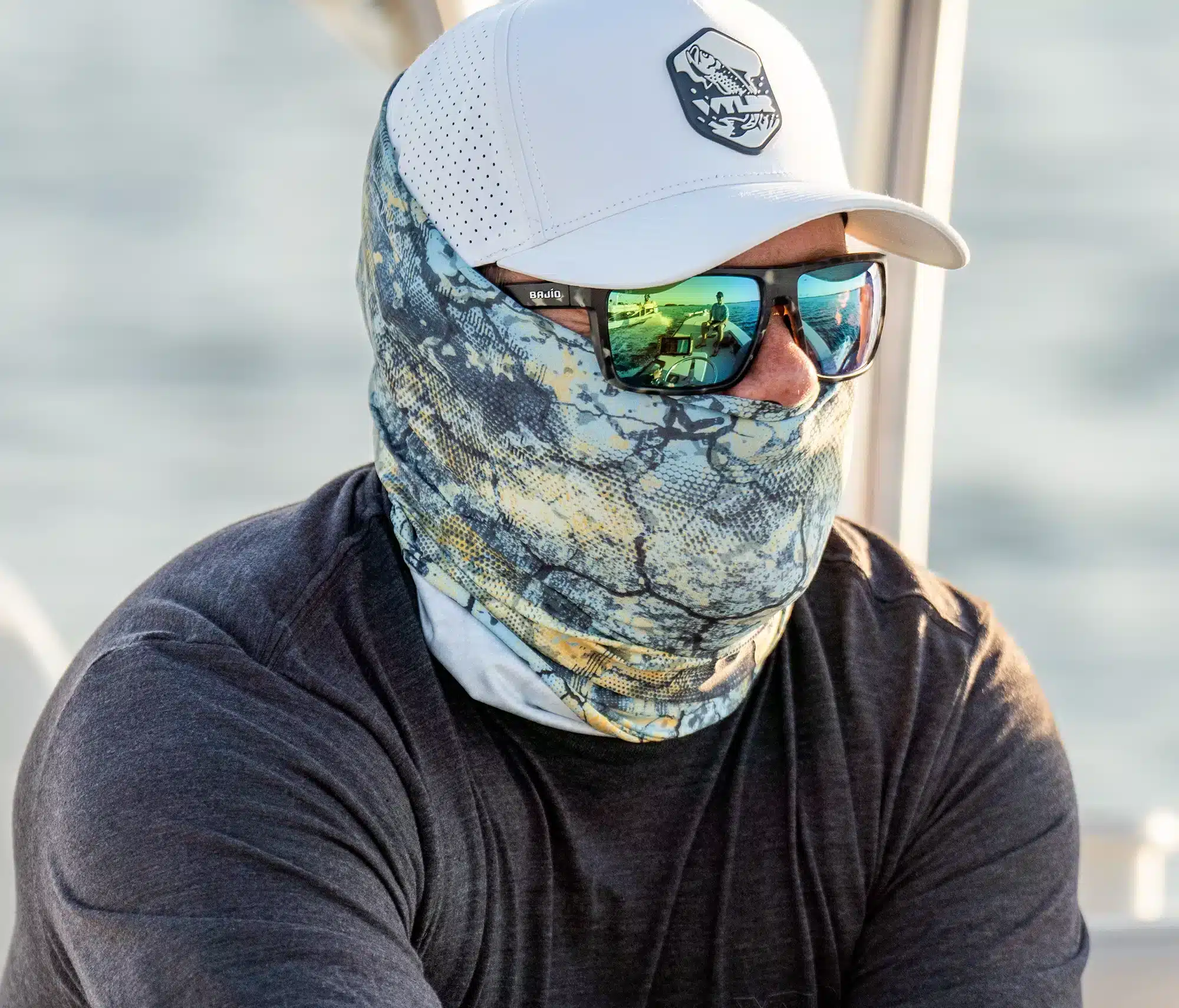 An image of a person driving a boat. They are in an off-white colored hat, a neck gaiter, sunglasses, and a charcoal black shirt. The hat features a tarpon fish breaching water. You can see the passengers in the reflection of the sunglasses.